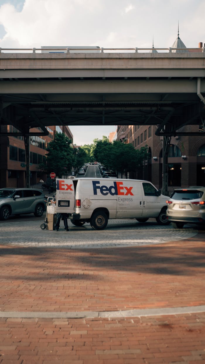 FedEx delivery van parked under an urban bridge in a busy city street scene with cars.