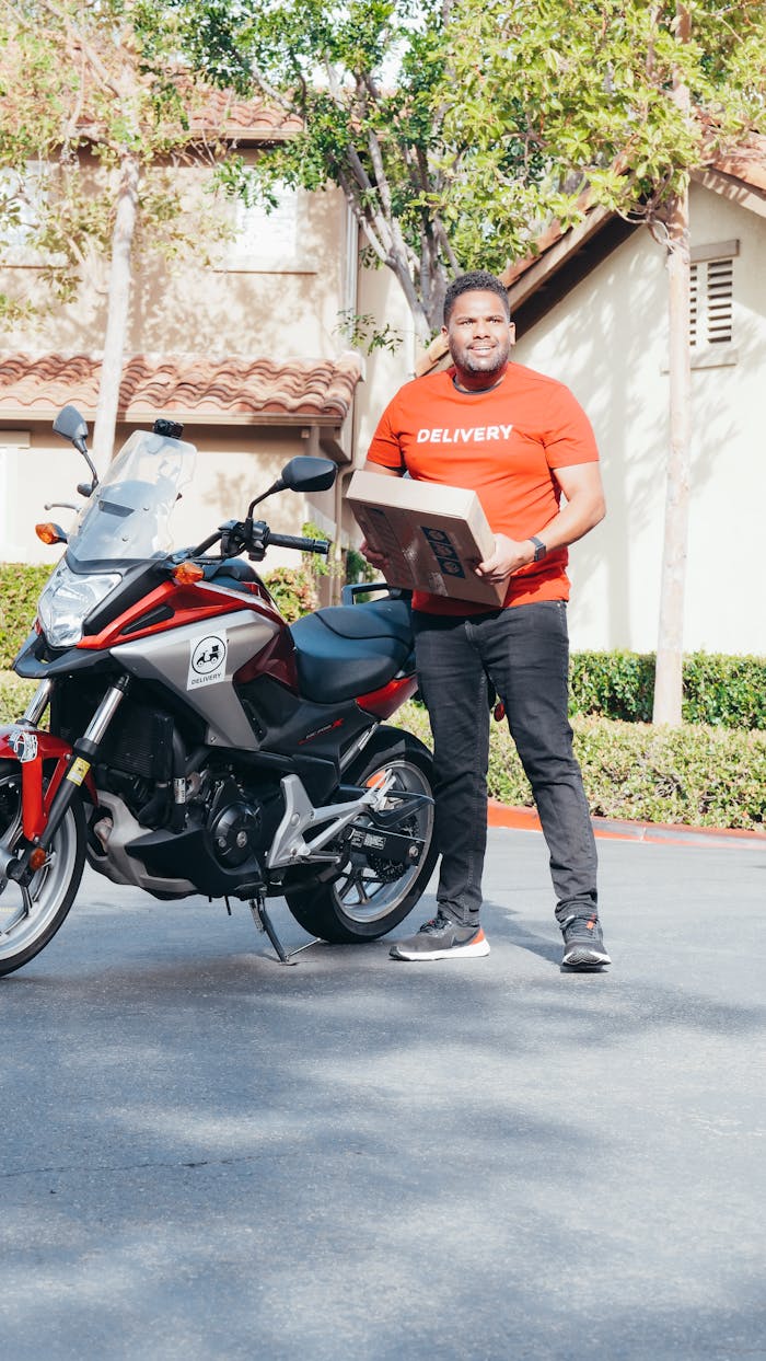 A delivery man in a red uniform holding a package beside a motorcycle in an outdoor setting.
